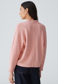 Rosa, weicher Pullover mit lockerer Passform und überschnittenen Schultern, mit einem hohen Halsausschnitt und feiner Textur. Kombiniert mit dunklen Jeans.