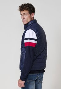 Chaleco acolchado azul marino con cuello alto, cierre frontal y bolsillo lateral. Presenta mangas a rayas en blanco, rojo y azul marino sobre una camiseta oscura debajo.