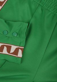 Gröna tygshorts med ett strukturerat randmönster i brunt, med matchande gröna knappar på sidofickan och elastisk midja.