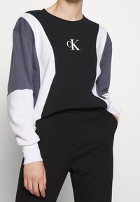 Sudadera negra con bloques de color blanco y gris, con un cuello redondo y un logotipo CK en contraste en la parte delantera. Tejido suave y texturizado.