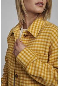 Žlutá a bílá kostkovaná vlněná bunda s límcem, velkým knoflíkem a kapsou na prsou. Měkká textura, viditelné detaily šití.