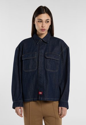 Femme portant une veste en jean bleu foncé oversize avec deux poches poitrine et un pantalon beige, debout devant un fond clair uni.