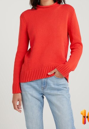 Frau trägt einen knallroten, gestrickten Pullover und hellblaue Jeans, mit einer Hand in der Tasche vor einem schlichten, hellen Hintergrund.