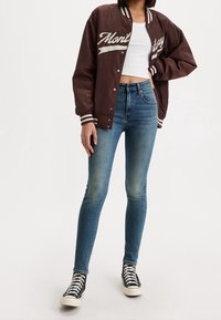 Brun bomberjacka med vit text "Monterey", vita ribbade manschetter, kombinerad med bleka blå skinny jeans och svarta högskor.