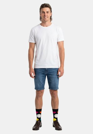Homme debout, portant un t-shirt blanc, un short en denim, des chaussettes noires avec des formes colorées, et des chaussures en cuir marron, sur un fond uni.