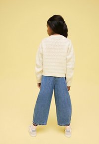 Pulover alb tricotat cu un model texturat, asortat cu blugi tocmai largi de culoare albastră. Copilul poartă adidași de culoare deschisă, văzut din spate.