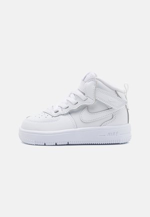 Witte high-top Nike sneaker met veters, een klittenbandsluiting en een gestructureerde zool op een lichtgrijze achtergrond.