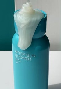 En turkis plastflaske merket "AFTER SUN SHOWER GEL" med en kremet hvit gel som renner fra toppen. Glatt tekstur og minimalistisk design.