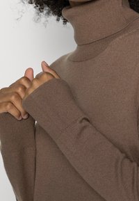 Maglione a collo alto marrone realizzato in morbido tessuto a maglia, con maniche lunghe e polsini a coste, e un design aderente attorno al collo.