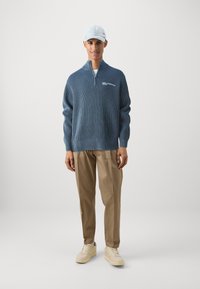 Pulover albastru cu nervuri, cu fermoar la jumătate, logo brodat, asortat cu pantaloni bej deschis și pantofi crem, completat de o șapcă palidă.