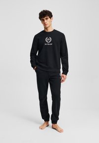 Hanorac negru și pantaloni jogger asortați, confecționați dintr-un material moale. Hanoracul are un logo alb și broderie decorativă pe piept.