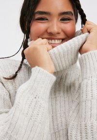 Hellgrauer Strickpullover mit hohem Kragen, gerippter Textur und dicken Bündchen. Der Pullover fällt weich und wirkt gemütlich.