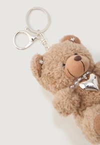 Porte-clé en forme d'ours en peluche brun et moelleux avec un pendentif en forme de cœur argenté, des accents en strass sur les oreilles et un fermoir en argent sur un fond blanc.