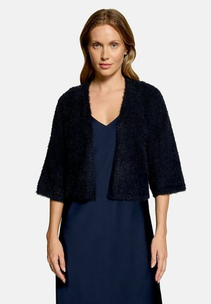 Woman with light brown hair wearing a dark fuzzy open-front cardigan over a sleeveless navy blue dress, standing against a plain light background.