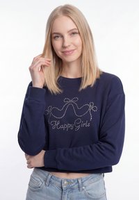 Fiatal nő egyenes szőke hajjal, söt�étkék pulóvert visel, melyen strasszköves masnik és "Happy Girls" felirat látható, világoskék farmerral párosítva.