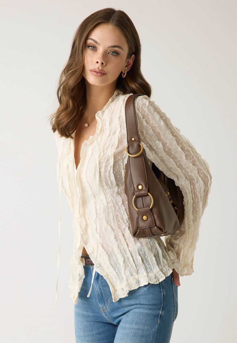 Femme aux cheveux bruns ondulés portant un chemisier crème transparent et un jean bleu, avec un sac à bandoulière en cuir marron, debout devant un fond uni.