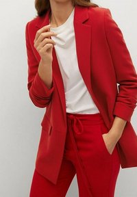 Blazer rouge à coupe ajustée avec revers crantés, associé à un pantalon rouge à taille élastique et un t-shirt blanc. Texture de tissu lisse partout.
