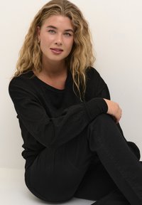 Sort, struktureret sweater med afslappet pasform og bred halsudskæring, parret med sorte bukser. Håret er bølget og indrammer ansigtet.
