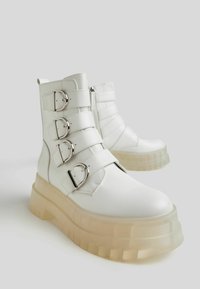 White leather ankle boots with four adjustable straps and silver buckles. Features a chunky, transparent platform sole with a textured finish.