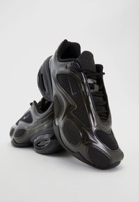 Paire de baskets Nike noires avec panneaux en mesh et synthétiques, design à lacets et semelles épaisses rembourrées sur un fond clair uni.