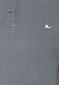 Polo gris texturé avec patte à deux boutons et petit logo blanc brodé sur la poitrine gauche indiquant "Petrol."