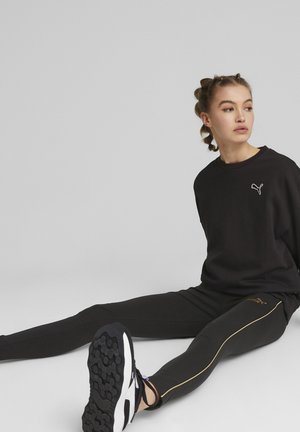 Czarna bluza z białym logo, czarne legginsy ze złotym paskiem po boku, w połączeniu z czarno-białymi butami sportowymi. Gładka tekstura, luźny krój.