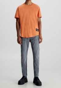 Orange bomullströja med korta ärmar, rund halsringning och en svart logopatch, kombinerad med grå slim-fit jeans och svarta klumpiga skor.