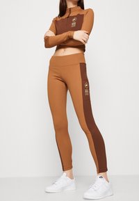 Hnědá sportovní souprava s dlouhým rukávem, obsahující crop top se zipem a odpovídající legíny s vyšším pasem, které mají barevné blokování a boční kapsu.