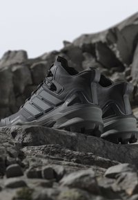 Chaussures de randonnée noires avec une finition texturée, dotées de trois accents de sangle, d'un col haut et de semelles robustes. Posées sur un terrain rocheux.