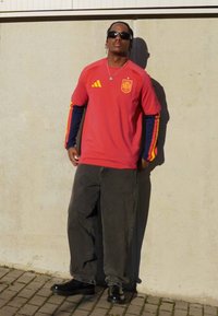 Rood Adidas-shirt met gele en blauwe strepen, met een wapenschild van Spanje. Draagt een donkergrijze loszittende broek en zwarte schoenen.