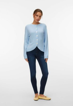 Femme portant un cardigan boutonné bleu clair, un jean skinny foncé et des ballerines jaune pâle, debout devant un fond blanc uni.