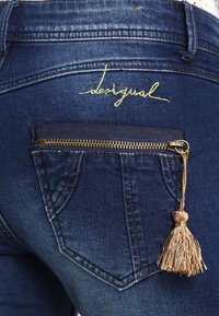 Jean en denim bleu foncé avec un logo "desigual" brodé en or et une poche zippée avec un détail de pompon doré.