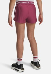 Bordeauxkleurige sportshorts met een brede witte tailleband met het label "UNDER ARMOUR." De stof is glad en heeft zijstrepen. Zwarte sportschoenen met witte zolen.