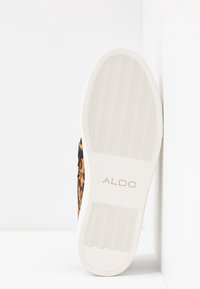 Semelle d’une chaussure en caoutchouc blanche avec un motif ondulé et le nom de la marque ALDO, montrant partiellement la tige imprimée léopard marron et noir.