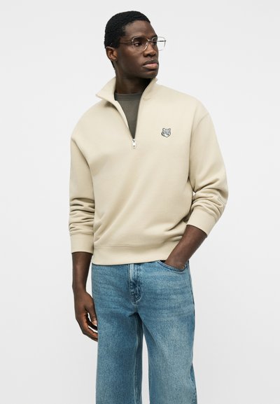 Άνδρας που φοράει μπεζ φούτερ με φερμουάρ μέχρι τη μέση και πατς λύκου, μπλε τζιν και γυαλιά, στέκεται με το ένα χέρι στην τσέπη και κοιτάζει προς τα δεξιά.