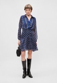 Mėlynos raštuotos suknelės su apykakle, permatomomis rankovėmis ir klostėmis. Sujungta su kelienų ilgio juodais batais ir tekstūruota juoda rankine.