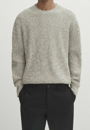 Homme portant un pull en maille texturée gris clair à manches longues et un pantalon noir, les mains dans les poches, debout devant un fond uni.