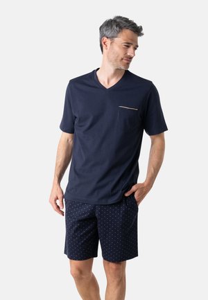 Homme portant un T-shirt bleu marine à col en V avec une poche poitrine et un short bleu marine à petit motif à pois, se tenant avec une main dans la poche.