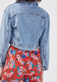 Veste en denim bleu clair à coupe courte, texture délavée et manches longues. Comprend deux poches avant à boutons et des surpiqûres décoratives.