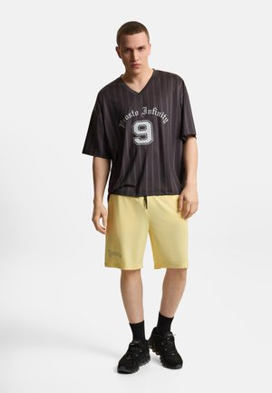 Mężczyzna w czarnym koszulce w paski z dekoltem w szpic z napisem „Prosto Infinity 9”, żółte spodenki z logo „Prosto”, czarne skarpety i czarne trampki.