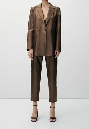 Vrouw gekleed in een bruin leren pak met een colbert met één knoop en cropped broek, gecombineerd met zwarte hoge sandalen met enkelbandje.