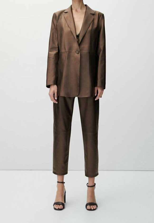 Femme portant un costume en cuir marron avec un blazer à un seul bouton et un pantalon court, associé à des sandales à talons noires avec bride à la cheville.