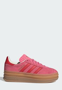 Sneaker de camurça rosa com detalhes em vermelho, apresentando três riscas e uma sola de borracha texturizada. A língua tem um padrão elevado.