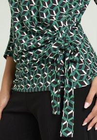 Groen en zwart topje met patroon en geometrisch ontwerp, voorzien van een knoopdetail aan de zijkant, gemaakt van rekbare stof. Gecombineerd met nauwsluitende zwarte broek.