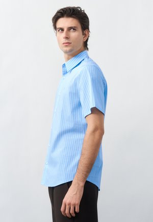 Νεαρός άνδρας που φοράει κοντομάνικο, ανοιχτό μπλε πουκάμισο με κάθετες ρίγες, στέκεται πλαγίως μπροστά σε απλό φόντο.