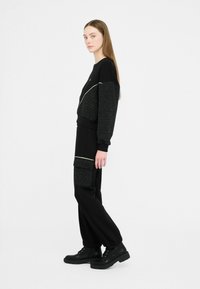 Zwarte sweatshirt met gestructureerde zwarte mouwen en licht accent, gecombineerd met losse zwarte broeken met een zakdetail. Draagt zwart laarzen.