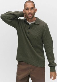 Khaki groene geribde trui met een polo kraag, drie zwarte knopen en lange mouwen. Draagt over een witte shirt, met een ontspannen pasvorm.