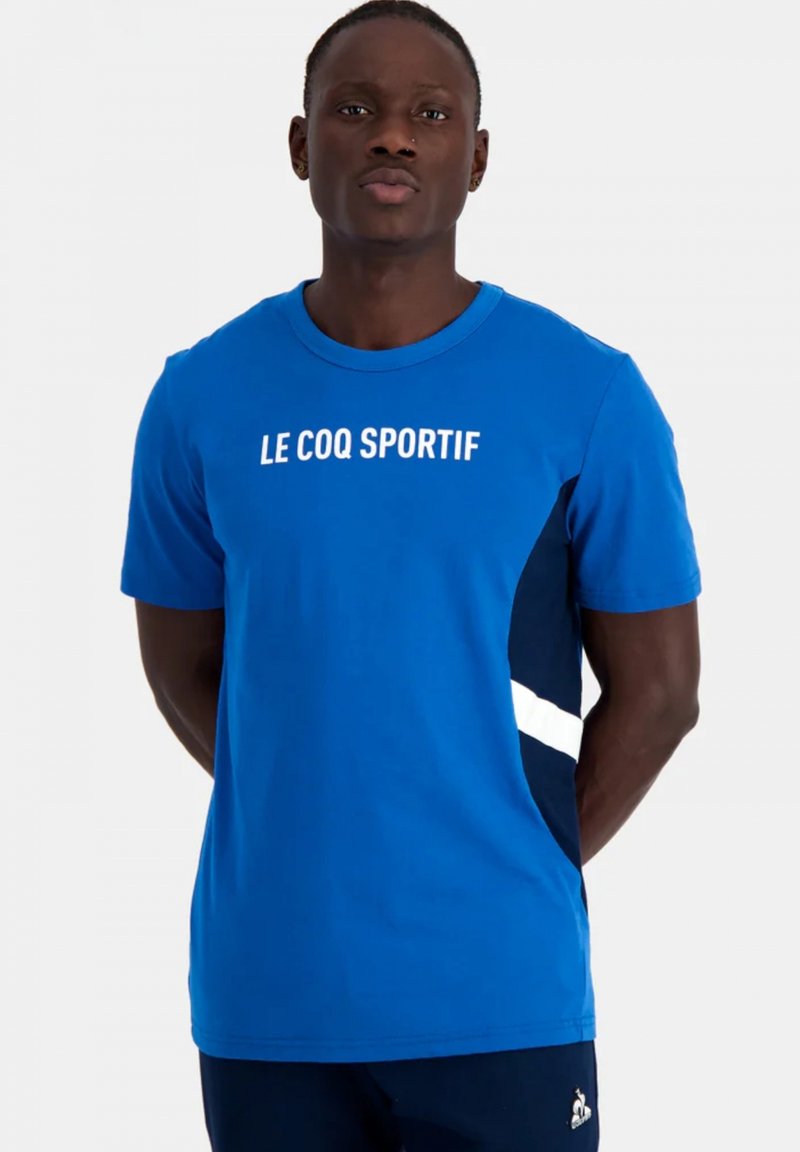 Camiseta de manga corta azul con un logo blanco de "LE COQ SPORTIF" en el pecho, con paneles laterales en color marino y diseño de cuello redondo.