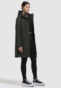 Mörkgrön lättviktsparka med huva, dragkedja framtill, fickor och avslappnad passform, bärs över svarta leggings och plattformsskor.