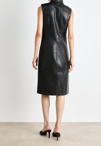 Zwarte lederen mouwloze jurk met een hoge kraag, een getailleerd silhouet en een naaddetail aan de achterkant, gecombineerd met open-toe zwarte hakken.